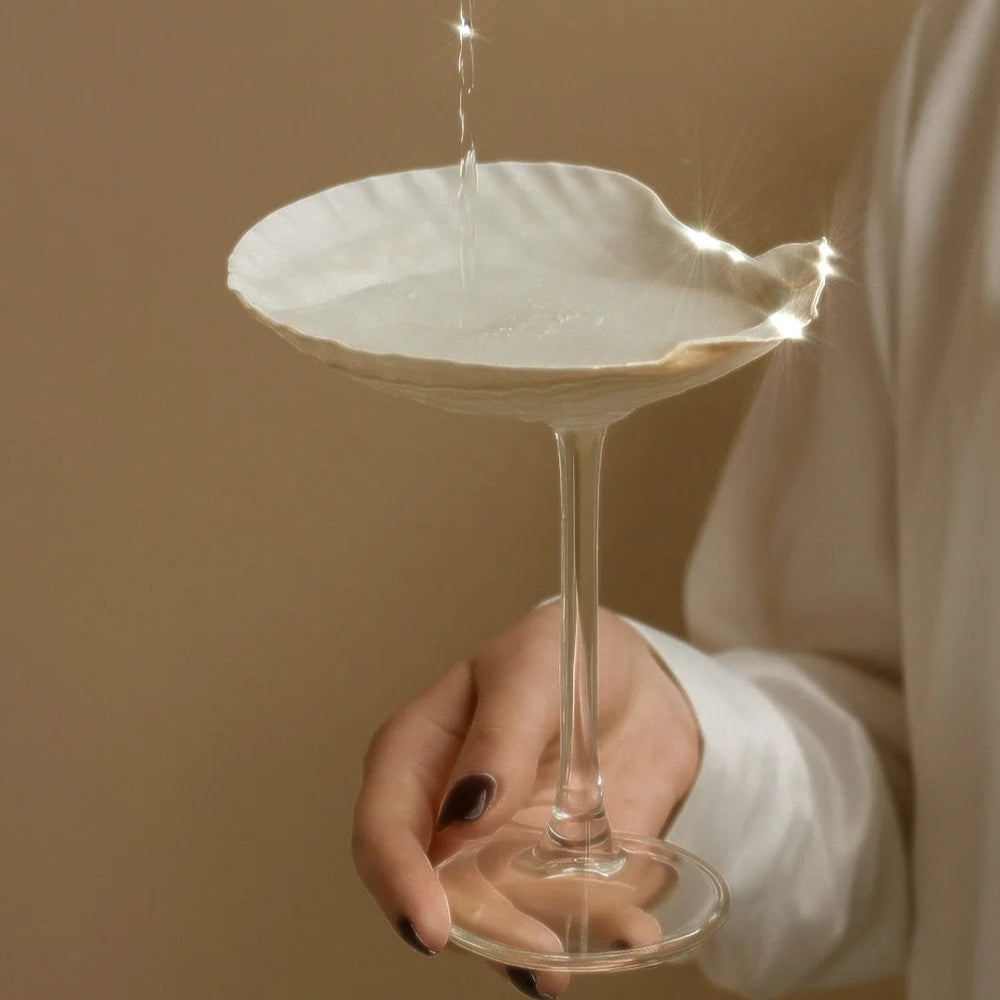 Person pouring liquid into a shell-shaped glass against a beige background