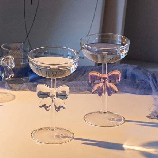 Two clear glass cocktail glasses with bow designs on a reflective surface.