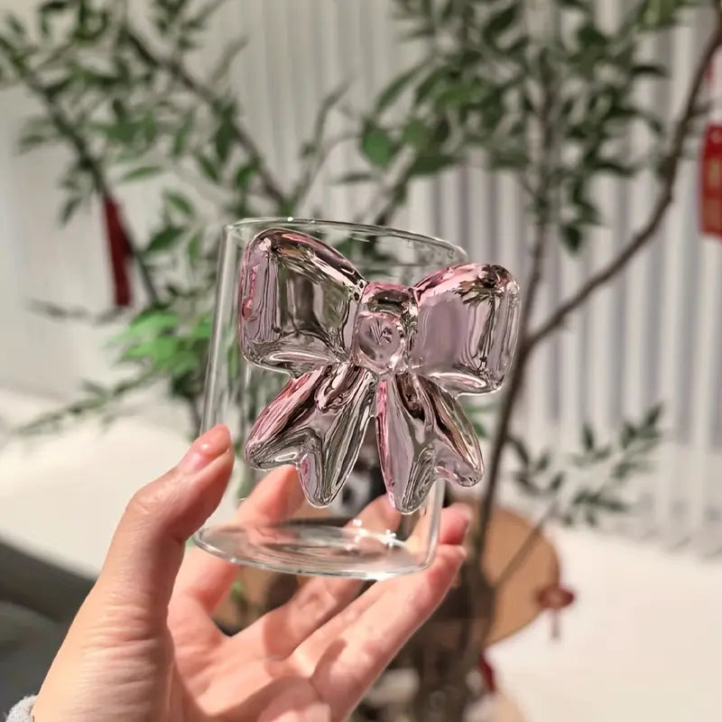 Clear glass mug with pink bow design held by a hand, blurred plant and indoor setting in the background