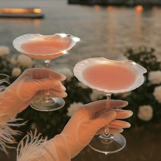 Two hands holding pink cocktails with a scenic lake and island in the background.