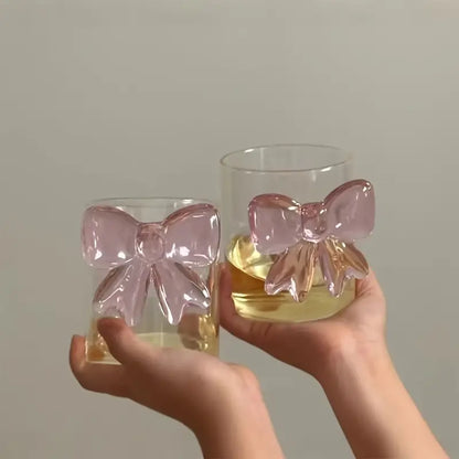 Two glasses with pink bow designs held by a hand against a neutral background
