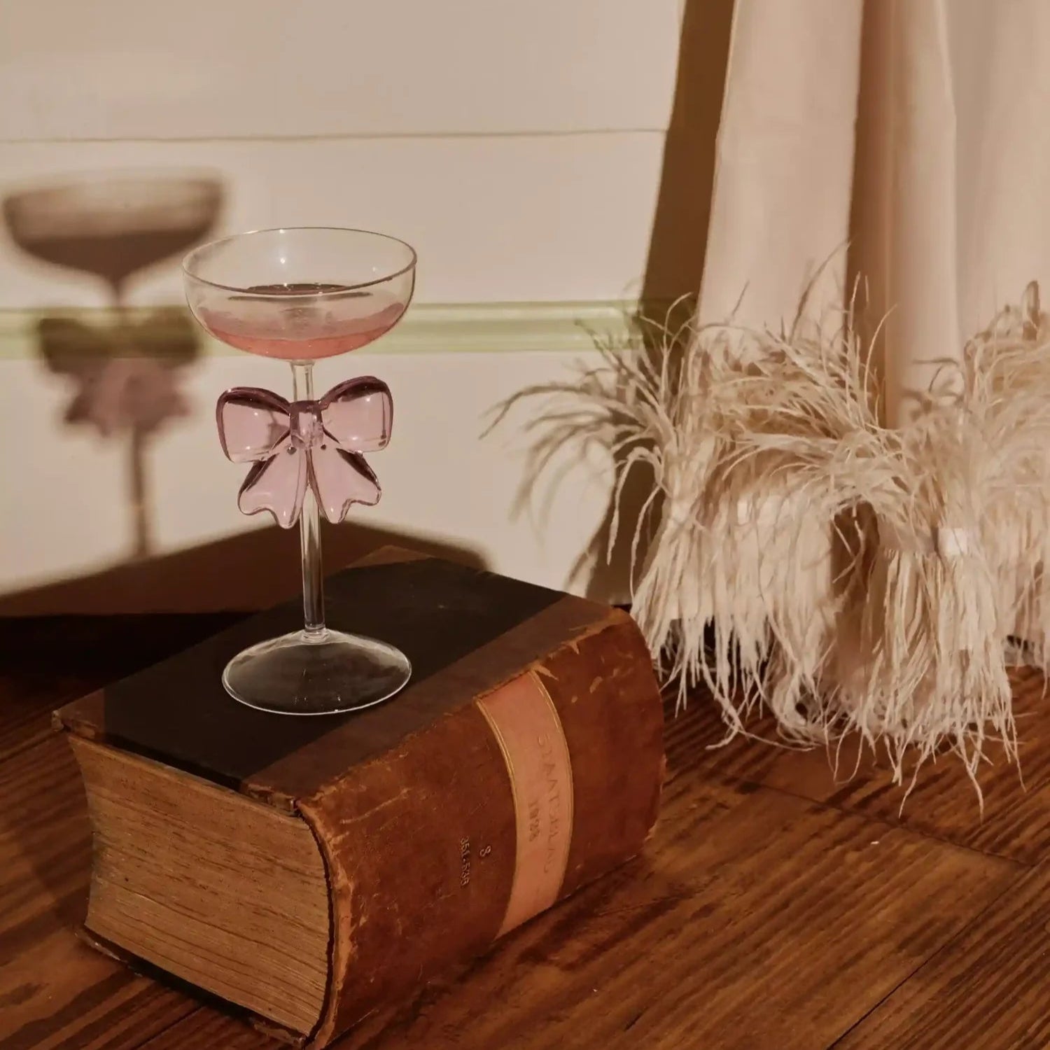 Cocktail glass with a pink drink and bow on an old book, with a plant in the background.